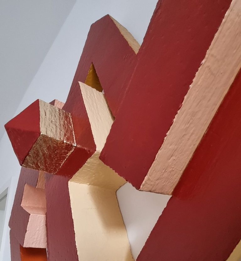 Sculpture abstraite en bois aux couleurs rouge et beige, avec des formes géométriques.